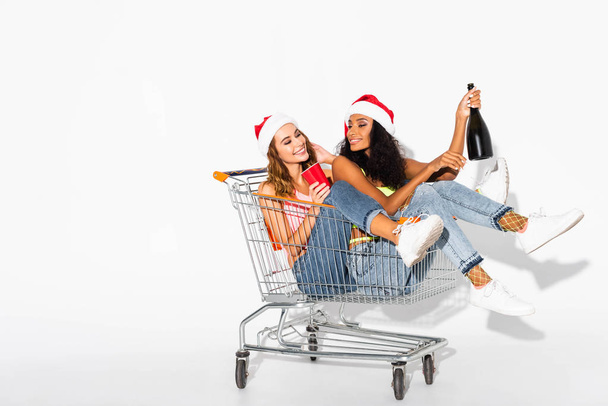 happy multicultural girls sitting in shopping cart with bottle of champagne on white  - 写真・画像