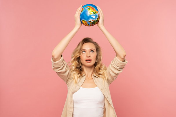Blonde woman holding globe above head isolated on pink - 写真・画像
