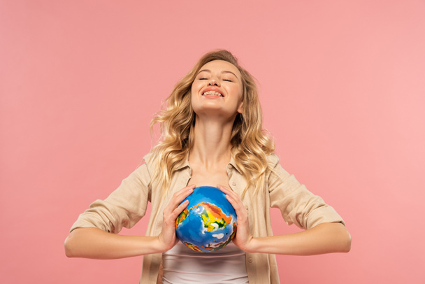 Smiling blonde woman holding globe isolated on pink - 写真・画像
