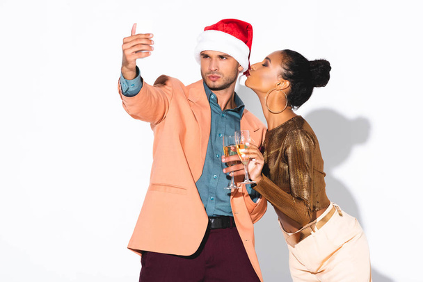 handsome man in santa hat taking selfie near african american girl with duck face on white  - 写真・画像