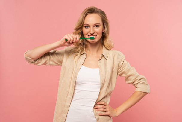 Smiling blonde woman holding toothbrush isolated on pink - 写真・画像