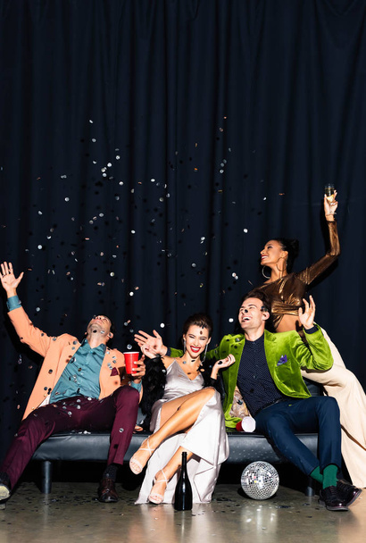 positive multicultural friends sitting on sofa near falling confetti on dark blue - Фото, изображение