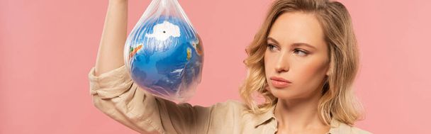 Panoramaaufnahme einer blonden Frau, die in einer Polyethylenpackung isoliert auf rosa, globalem Erwärmungskonzept auf den Globus blickt - Foto, Bild