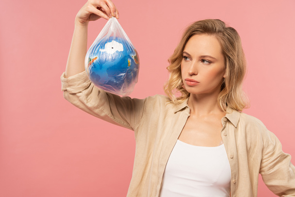 Pensive woman holding globe in polyethylene package isolated on pink, global warming concept - Photo, Image