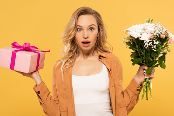 Shocked blonde woman holding bouquet and gift box isolated on yellow - Foto, Bild
