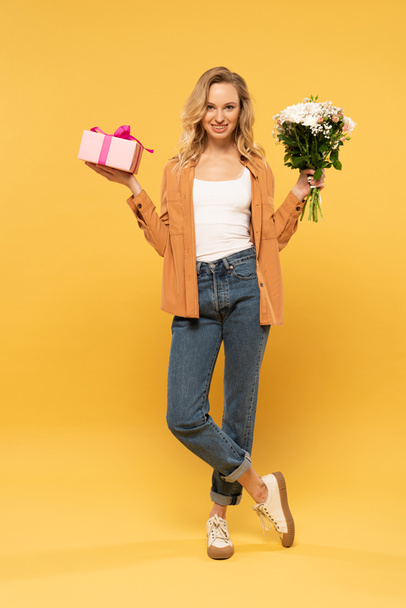 Smiling blonde woman holding bouquet and gift box on yellow background - Fotó, kép