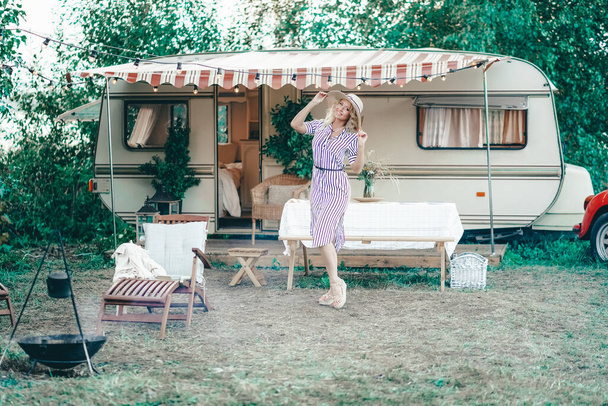 a cute woman with wavy blonde hair in a trendy summer striped dress on the background of a trailer camper van rests - Foto, imagen