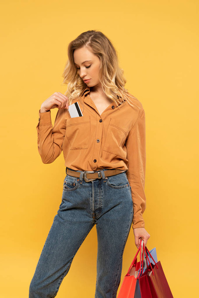 Blonde woman putting credit card in shirt pocket and holding shopping bags isolated on yellow - Foto, Imagen