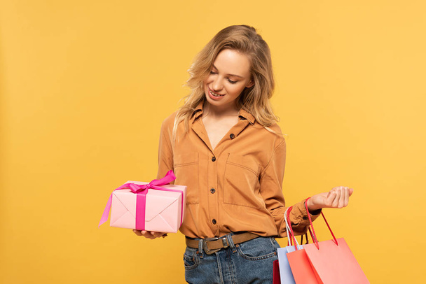 Smiling woman looking at gift box and holding shopping bags isolated on yellow - Фото, изображение