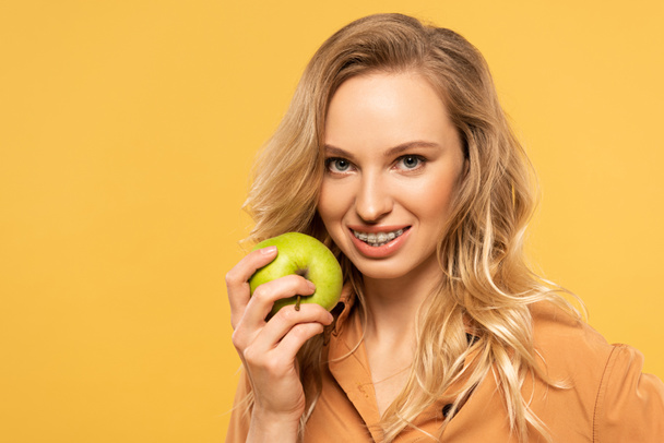 Χαμογελαστή ξανθιά γυναίκα με οδοντικά στηρίγματα κρατώντας πράσινο μήλο απομονωμένο στο κίτρινο - Φωτογραφία, εικόνα