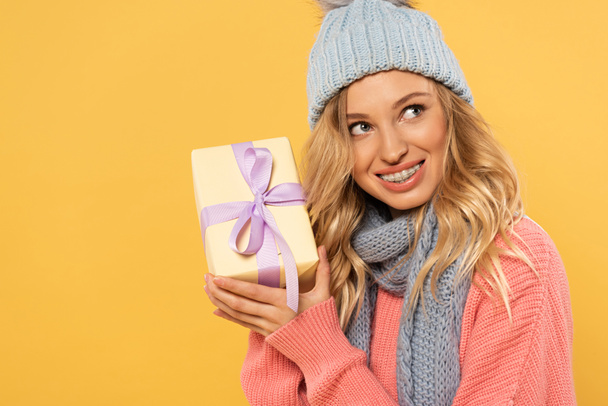 Smiling woman in hat and scarf holding gift box isolated on yellow - Fotó, kép