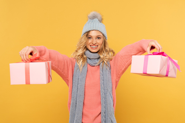 Smiling blonde woman in hat and scarf holding gift boxes isolated on yellow - Fotografie, Obrázek