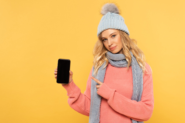 Woman in hat and scarf pointing on smartphone with blank screen isolated on yellow - Zdjęcie, obraz
