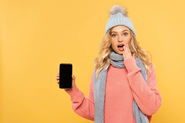 überraschte Frau mit Hut und Schal hält Smartphone mit leerem Bildschirm isoliert auf gelb - Foto, Bild