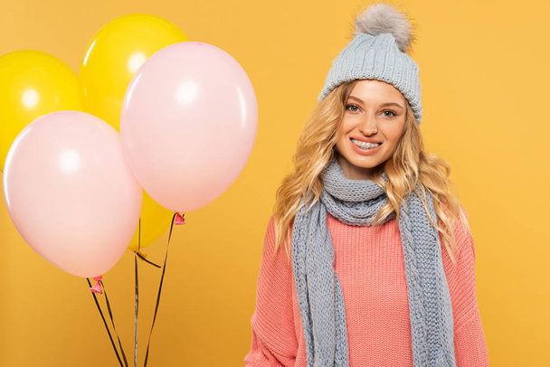 Woman in hat and scarf smiling and holding balloons isolated on yellow - Fotografie, Obrázek