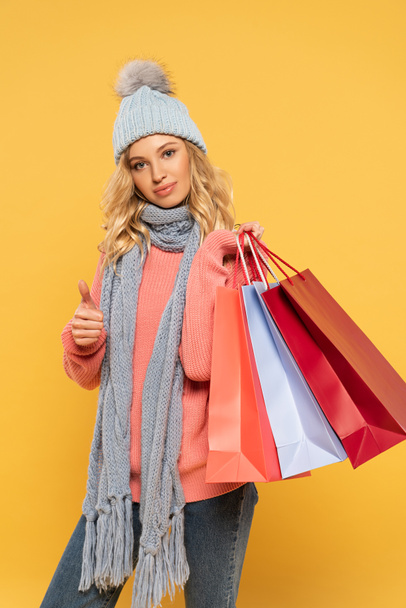Nő kalap és sál gazdaság bevásárló táskák és mutatja hüvelykujj felfelé jel elszigetelt sárga - Fotó, kép