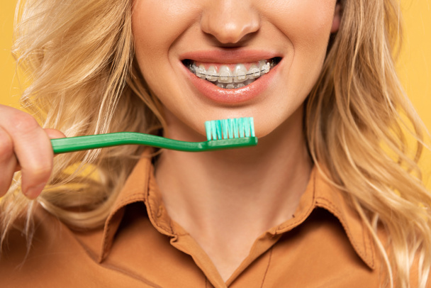 Oříznutý pohled na usmívající se ženu v zubní rovnátka držení zubního kartáčku izolované na žluté - Fotografie, Obrázek