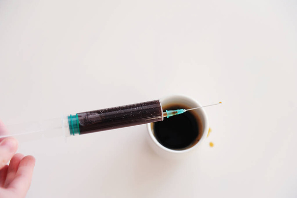 einen typischen Espresso in einer medizinischen Spritze in der Hand über einer Tasse. Symbol der Koffeinsucht. weißer Hintergrund isoliert - Foto, Bild