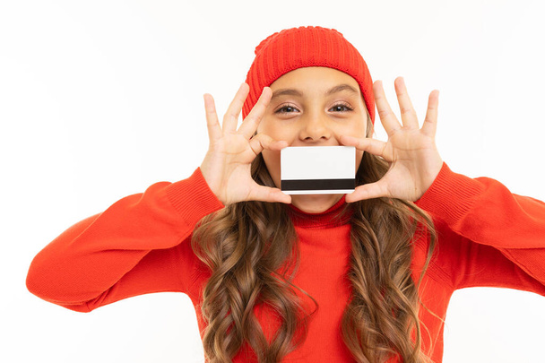 χαριτωμένο κοριτσάκι με κόκκινο πουλόβερ και καπέλο ποζάρουν με πιστωτική κάρτα σε λευκό φόντο  - Φωτογραφία, εικόνα