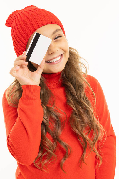linda niña en suéter rojo y sombrero posando con tarjeta de crédito sobre fondo blanco
  - Foto, Imagen