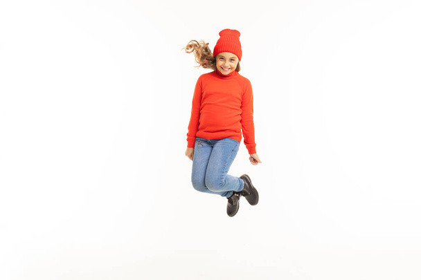 aranyos kislány piros pulóver és kalap jumping ellen fehér háttér  - Fotó, kép