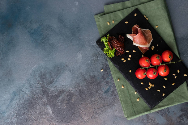 Prosciutto, cherry tomatoes and pine nuts. Fresh appetizer on the black board. Free space for text - Zdjęcie, obraz