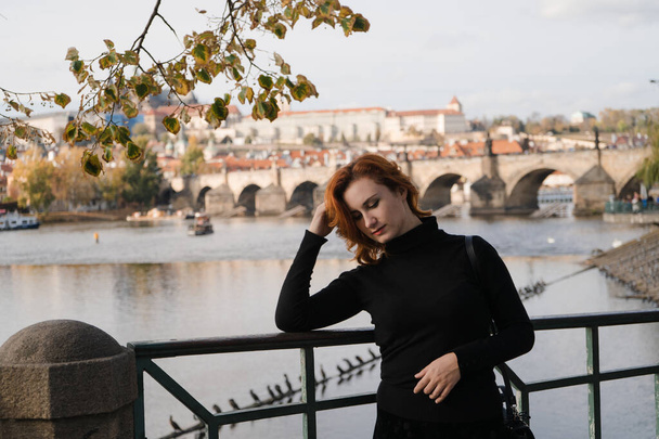 Happy redhead tourist traveler in Prague, Czech Republic wearing black sweater with Charles Bridge in the background - Photo, Image