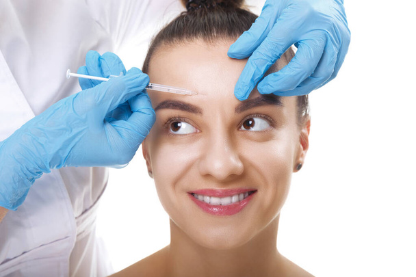 Egy fiatal, fehér, mosolygó nő portréja, aki kozmetikai botox injekciót kap a homlokába. Emberek, kozmetológia, plasztikai sebészet és szépség koncepció. - Fotó, kép