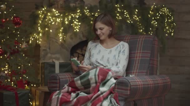 Young female in pajamas in armchair surfing in gadget and smiling with Christmas tree and blurred lights on fireplace behind in slow motion.  - Filmagem, Vídeo