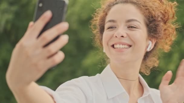 Charming happy young redhead woman in white shirt smiling and waving with hand while having a video call on smartphone in the park - 映像、動画
