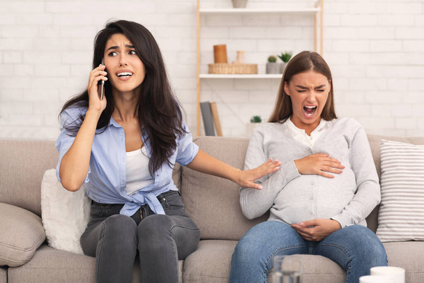 Menina chamando médico para amigo grávida ter espasmo sentado dentro de casa
 - Foto, Imagem