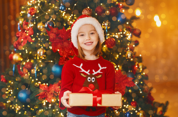 Κοριτσάκι που στέκεται δίπλα στο χριστουγεννιάτικο δέντρο κρατώντας το δώρο της - Φωτογραφία, εικόνα