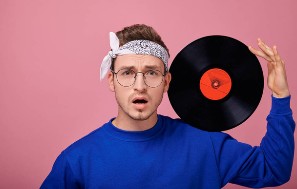 Cool stylish guy in white bandanna in dark blue sweater with vinyl record posing on pink background  - Fotografie, Obrázek