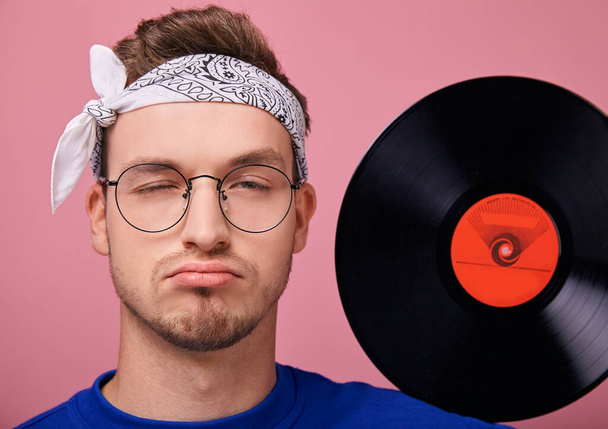 Cool stylish guy in white bandanna in dark blue sweater with vinyl record posing on pink background  - Foto, immagini