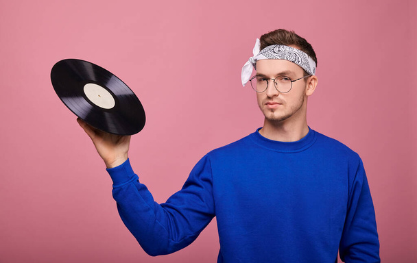 Cool stylish guy in white bandanna in dark blue sweater with vinyl record posing on pink background  - Fotó, kép