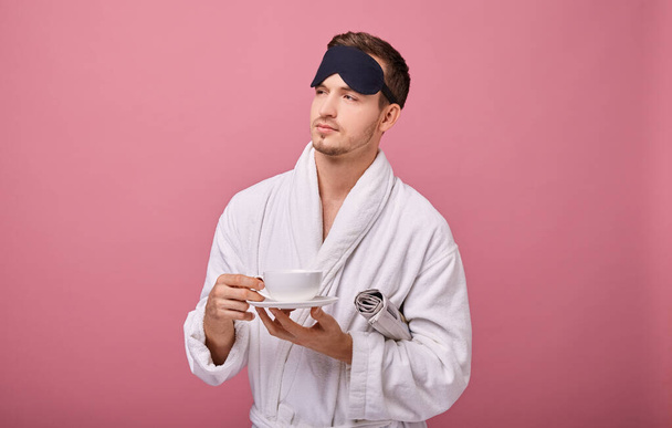 Un sleepy young man in sleeping mask and white coat with newspaper and cup of coffee on pink background - Fotoğraf, Görsel