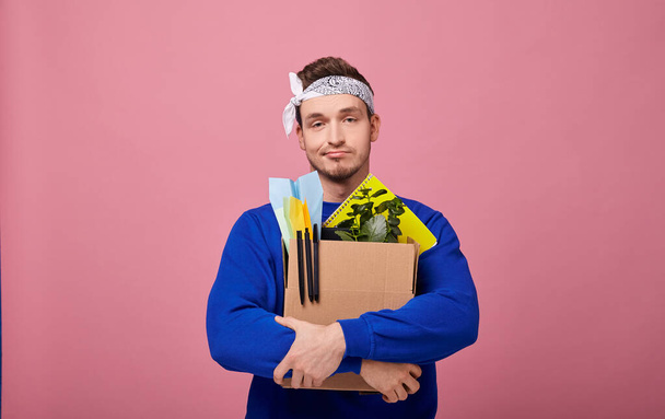 A cool stylish retro guy in a white with a bandana pattern in a dark blue sweater is standing hugging a box with both hands. The guy was fired from work. - Foto, Imagen