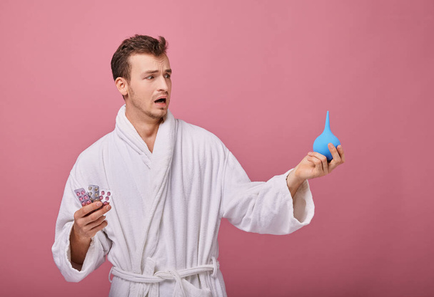 view of illness man standing on pink background holding enema and right pills  - Foto, imagen
