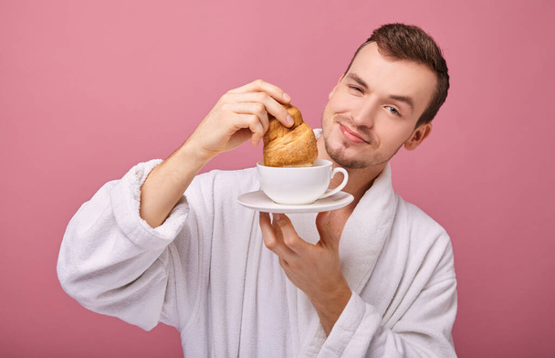 guy in white bathrobe standing with croissant in one hand and white cup on pink background  - Zdjęcie, obraz