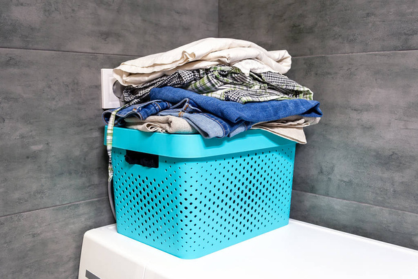 Cama dobrada, jeans, toalhas em uma caixa azul contra o fundo borrado de uma parede de concreto cinza no banheiro. Pilha de roupa e lavandaria no canto em uma máquina de lavar roupa
 - Foto, Imagem