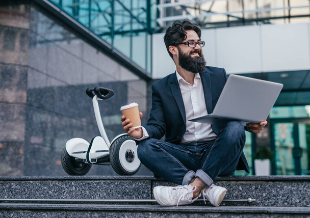 Cheerful manager with hot drink and laptop sitting on steps - Photo, Image