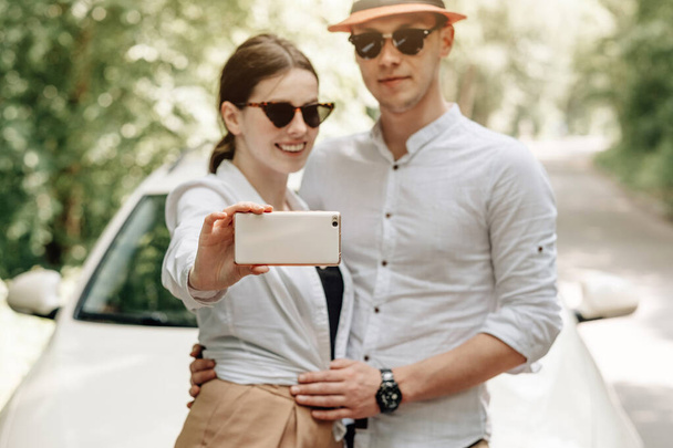 Young Happy Couple Dressed Alike in White T-shirt Holding Smartphone Near the Car, Weekend Outside the City, Holidays and Road Trip Concept - Фото, изображение