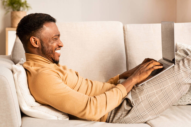 Happy African American Mies käyttäen kannettavaa tietokonetta makaa sohvalla sisätiloissa
 - Valokuva, kuva