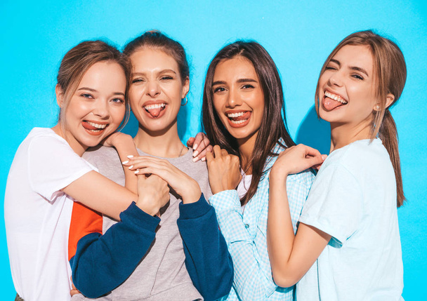 Quatro jovens lindas meninas hipster sorridentes em roupas de verão na moda. Mulheres despreocupadas Sexy posando perto da parede azul no estúdio. Modelos positivos se divertindo e abraçando. Eles mostram línguas
 - Foto, Imagem