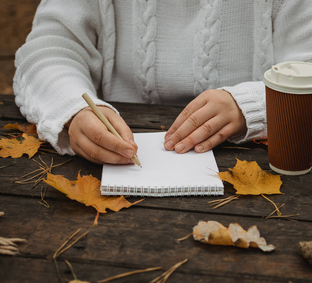 Emberi kezek egy csésze kávéval és sállal a fa asztalnál jegyzetfüzettel, tollal és őszi levelekkel. Emberi kéz egy csésze kávéval és sállal a fa asztalnál - Fotó, kép