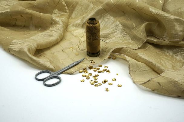 Gold sequins, spool of golden thread, needle on rippled gold silk background. Sewing and decorating accessories used for embellishment of dresses. - Photo, Image