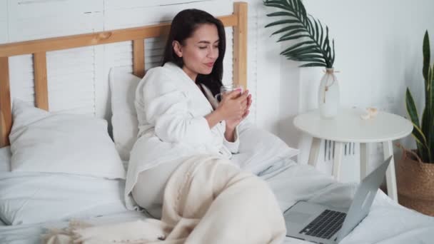 Woman in white bathrobe, lies on bed, drinks tea and watches movie on laptop - Video