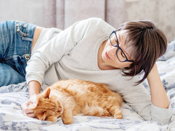 Cute ginger cat and woman in glasses are lying in bed. Woman is stroking her fluffy pet. Morning bedtime. - 写真・画像
