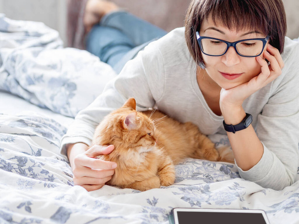 Cute ginger cat and woman in glasses are lying in bed. Woman is reading tablet and stroking her fluffy pet. Morning bedtime. - Valokuva, kuva