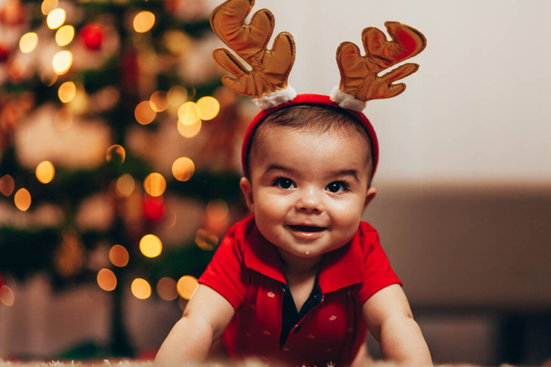 Cute baby boy wearing reindeer antlers crawling on floor over Christmas lights. Looking at camera. Holiday season. - Foto, afbeelding